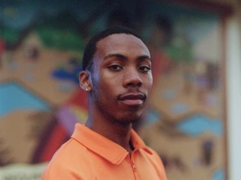 A portrait of Cipher Goings wearing an orange shirt and standing in front of a mural