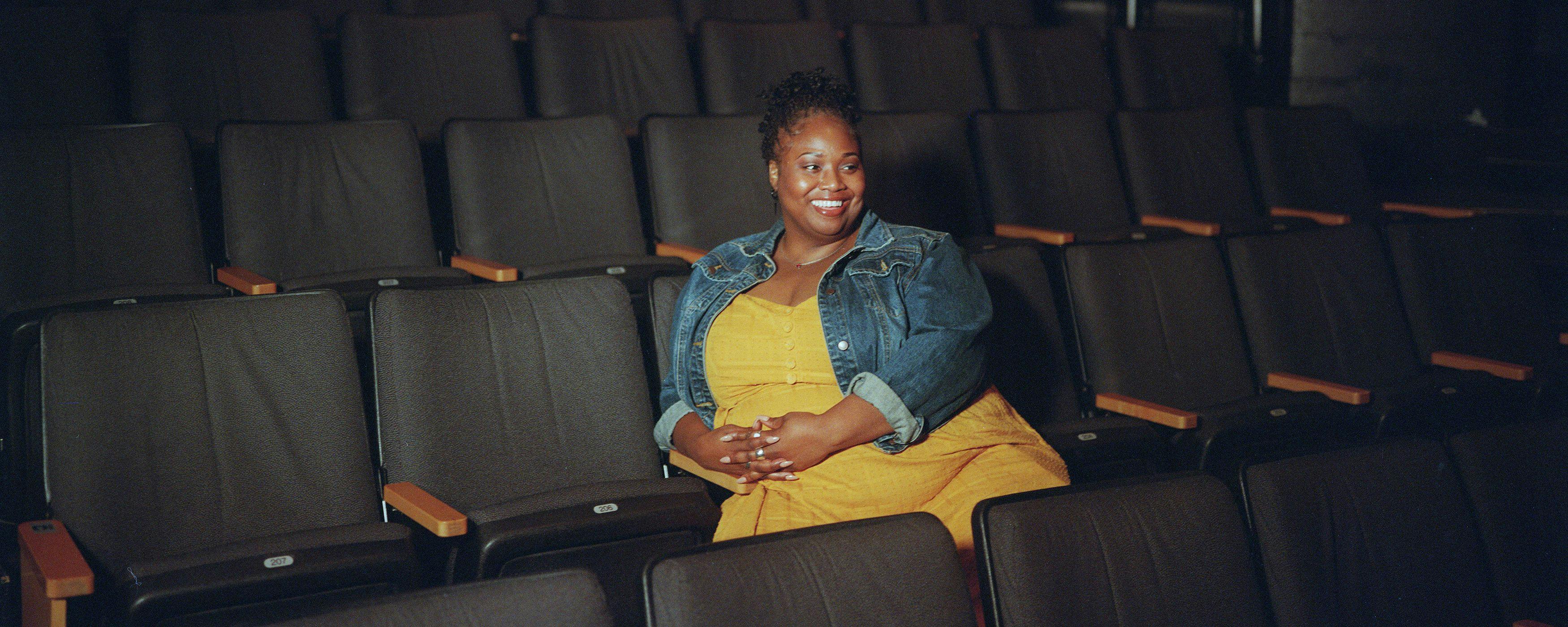 A smiling woman sits in a theater surrounded by empty seats. 