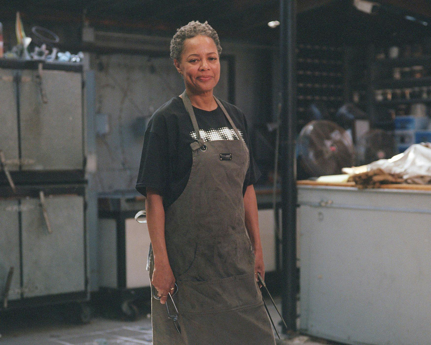 A woman with short grey hair wears a work apron in an industrial-looking art studio