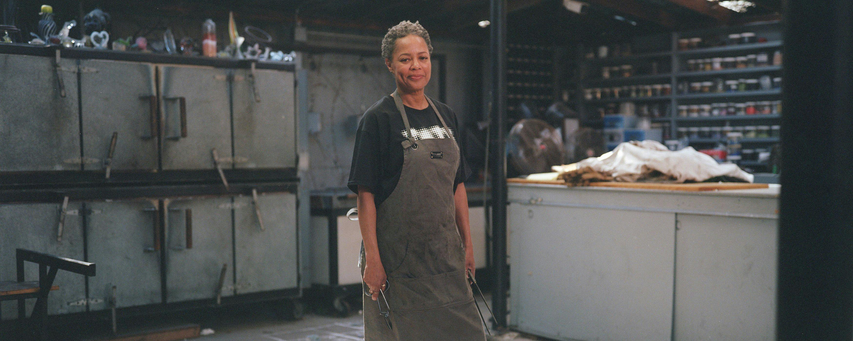 A woman with short grey hair wears a work apron in an industrial-looking art studio