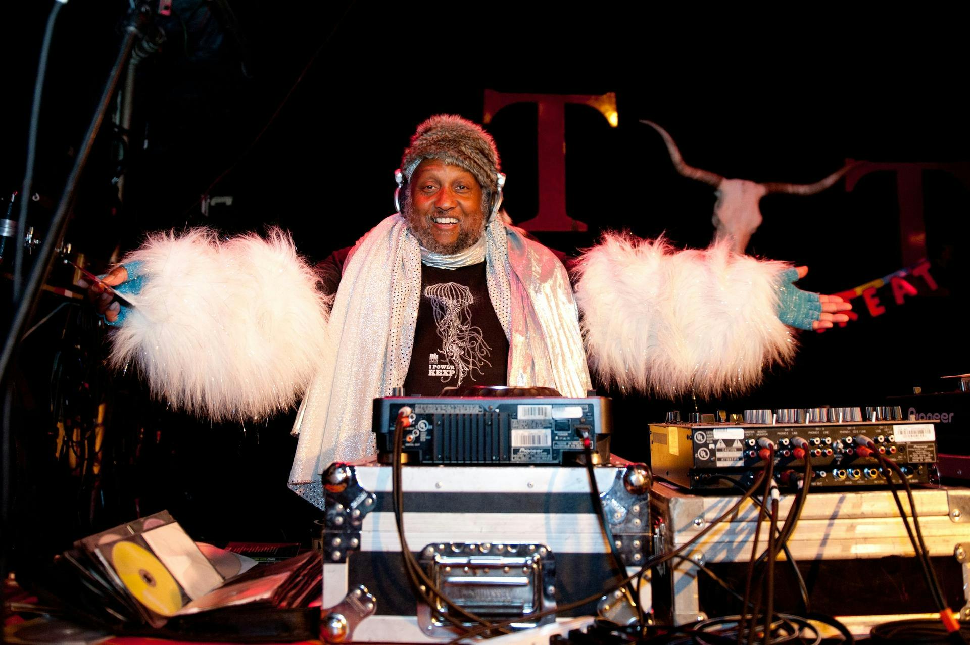 Riz Rollins stands behind DJ equipment wearing a sparkly silver scarf and white fuzzy sleeves