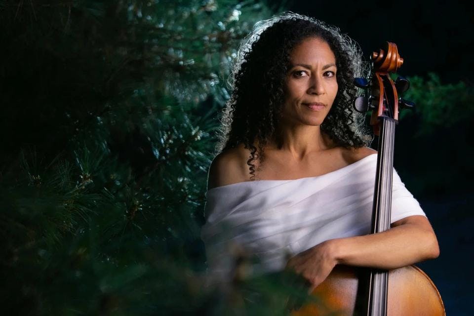 A portrait of Gretchen Yanover with her cello and an evergreen tree behind her
