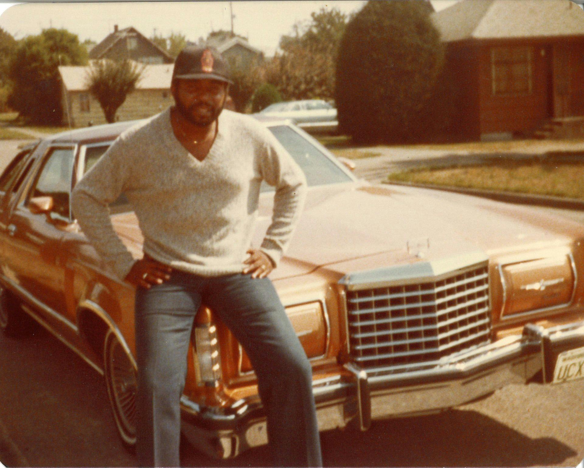 Robert L. Scott sits of the hood of a burnt umber-colored car wearing jeans, a v-neck sweater and a baseball cap