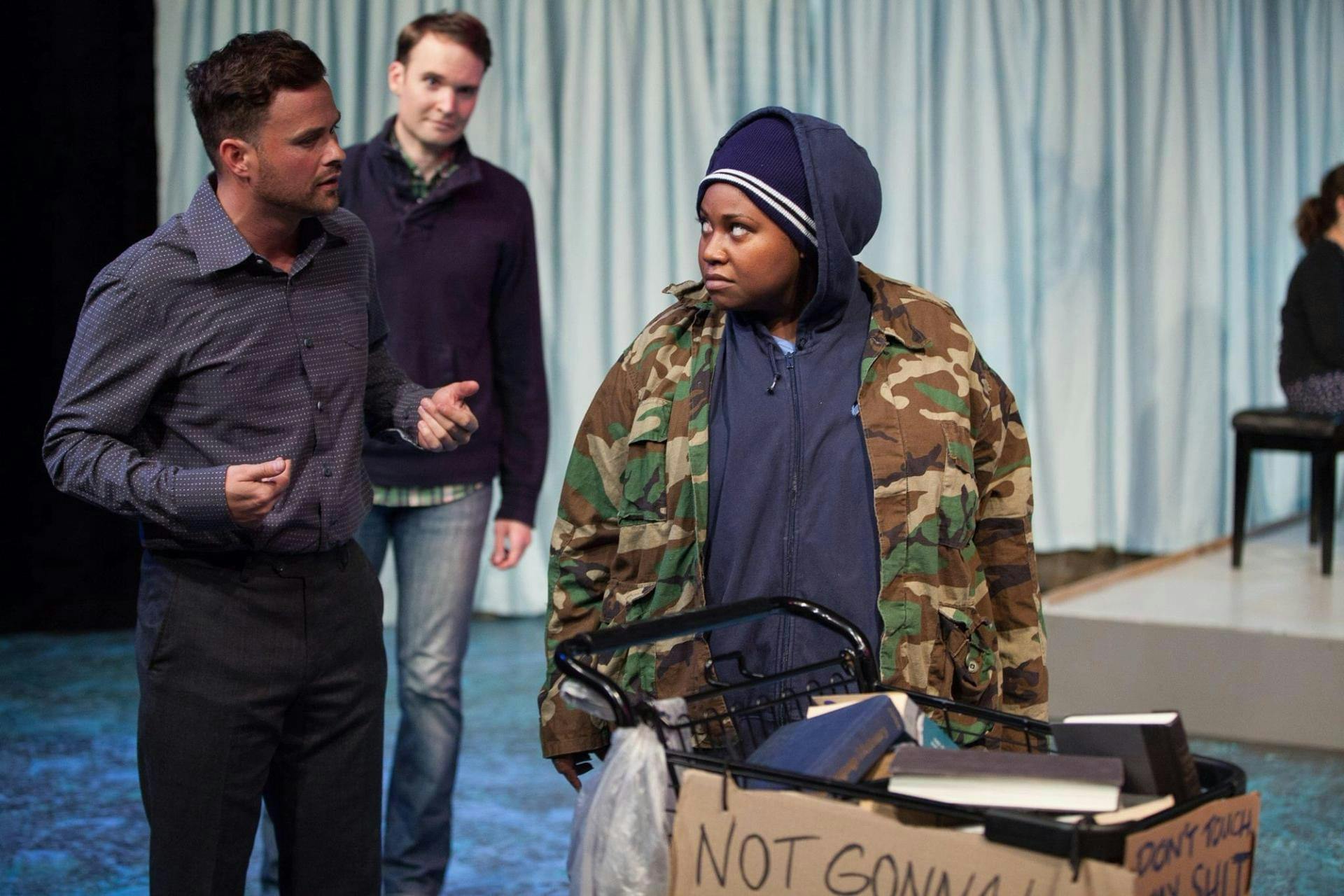 Three actors on stage, one of whom pushes a shopping cart