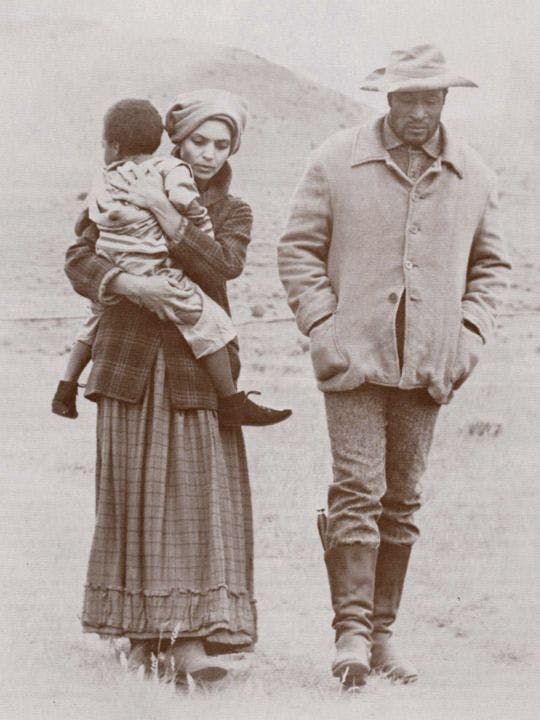 A film still in which a man and woman walk together in pioneer costumes; the woman holds a child