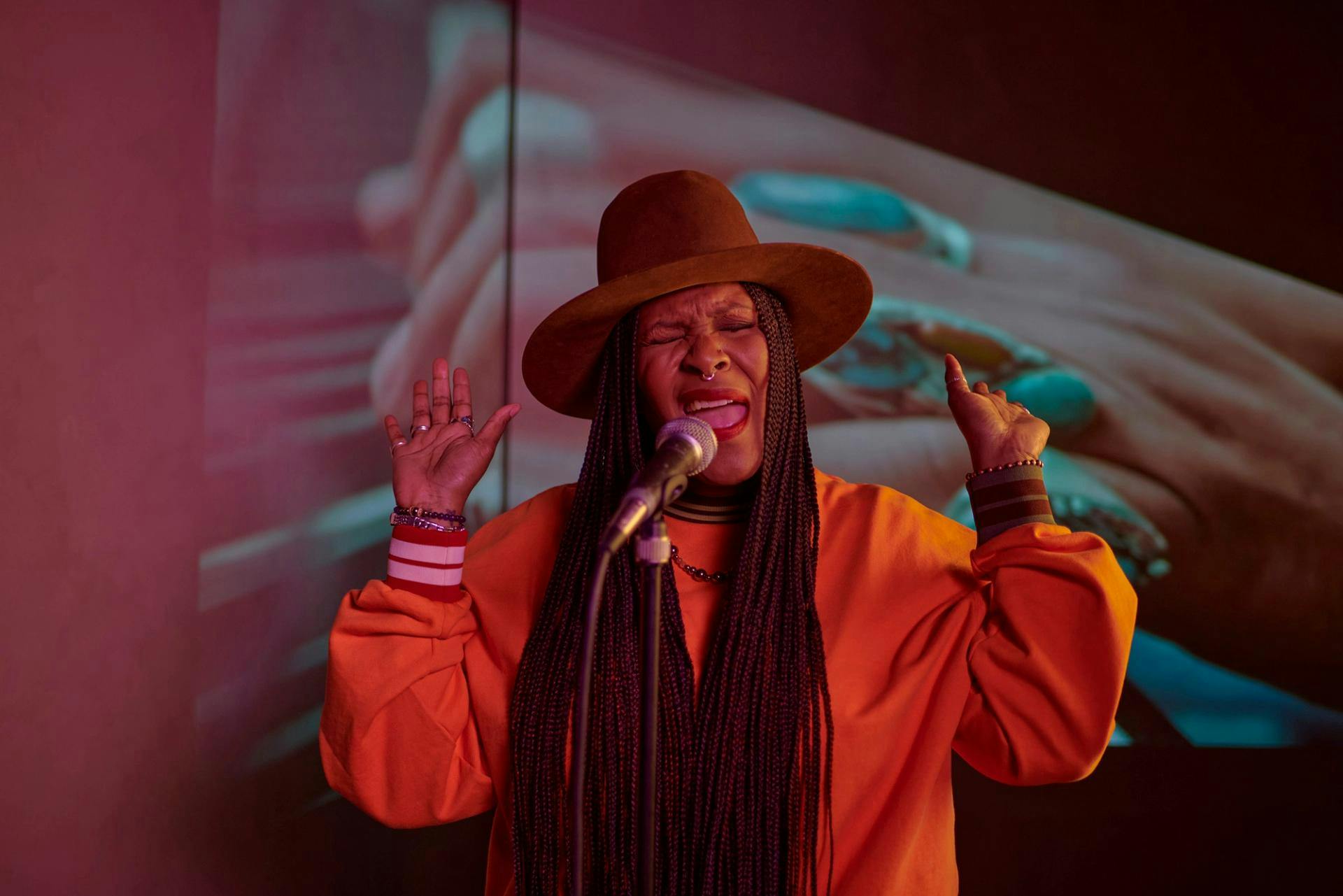 A woman wearing a hat sings into a mic passionately, her eyes closed and hands up