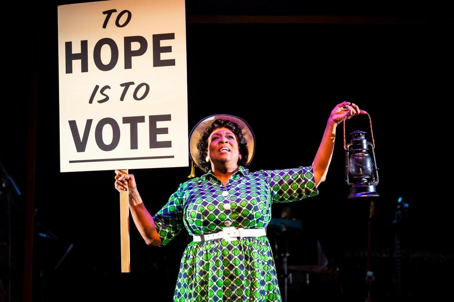 A woman in period costume holds a sign that reads "To hope is to vote" in one hand and a lantern in the other