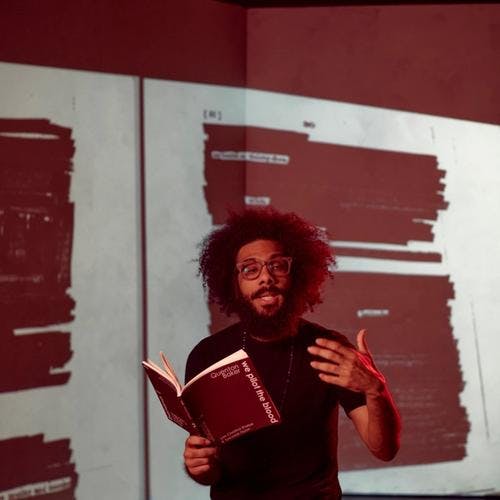 Person with blackout poetry behind them reads from a book, looking at the camera and gesturing with hand