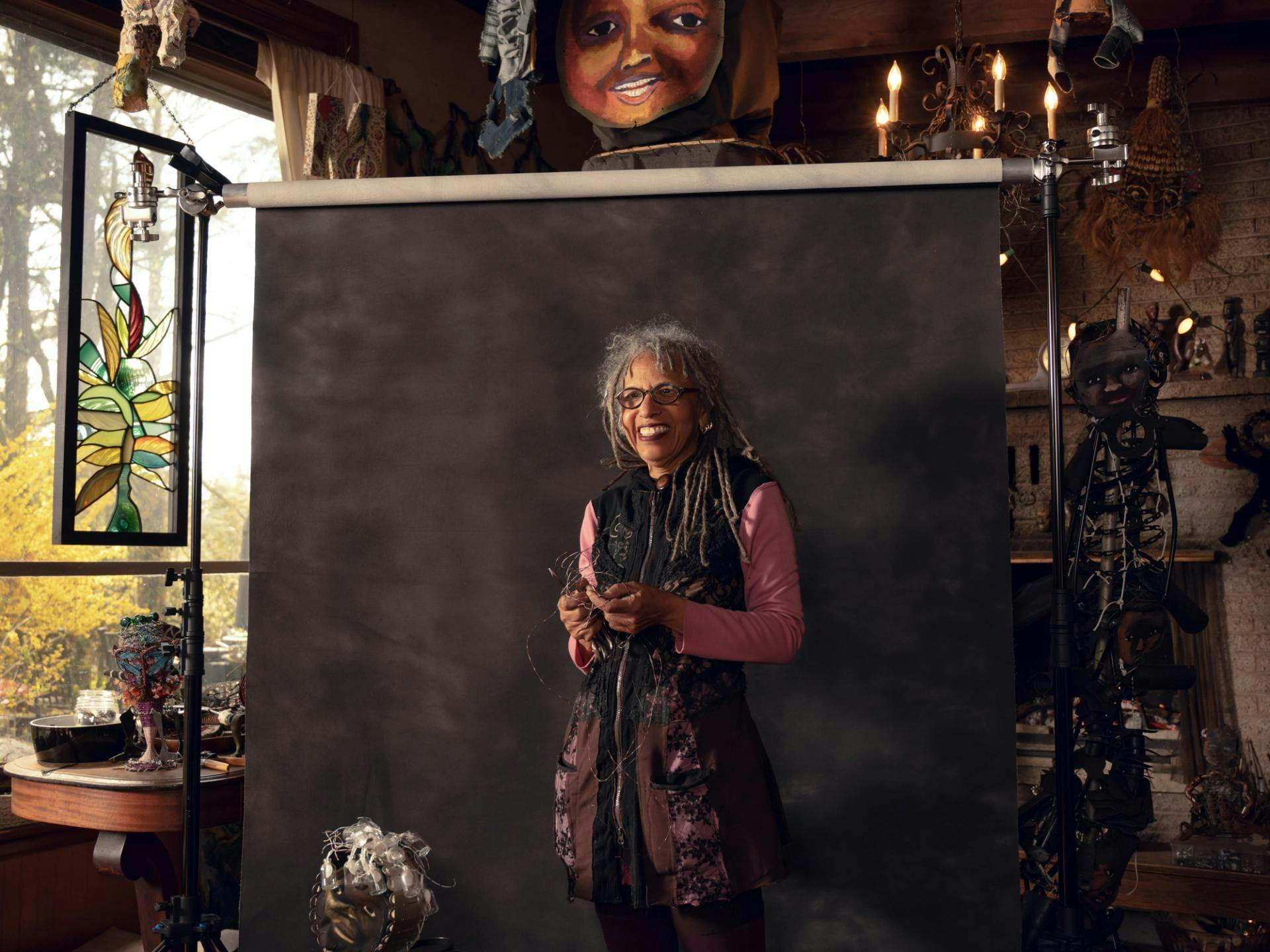 Person with glasses poses in front of a black backdrop in art studio full of objects