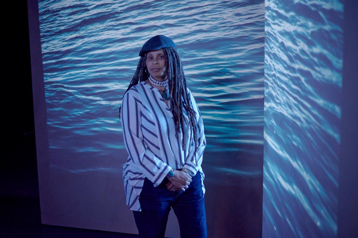 A woman stands in front of a projection of the ocean with her hands clasped