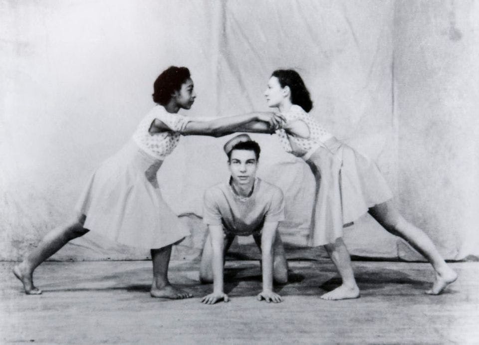 A black and white photo of two women holding each other's arms above a boy who is on all fours between them