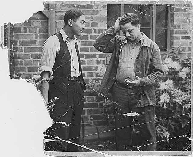 A torn vintage black and white photo of two men, one looking at the other and one looking down at his hand