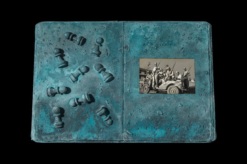 An open book painted blue with chess pieces on the left page and a black and white photo on the right page