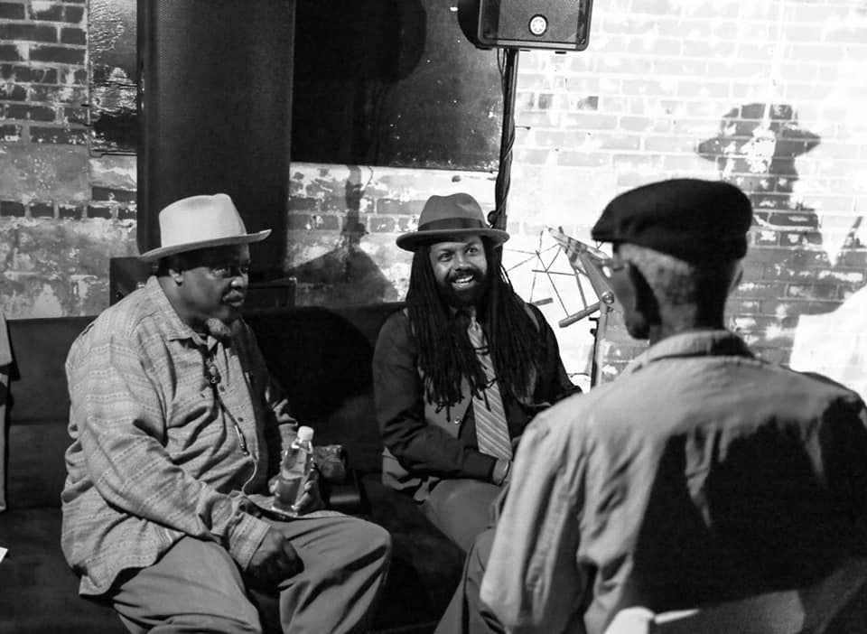 A black and white photo of Three men sitting together and talking; two men face the camera and the last has his back to the camera