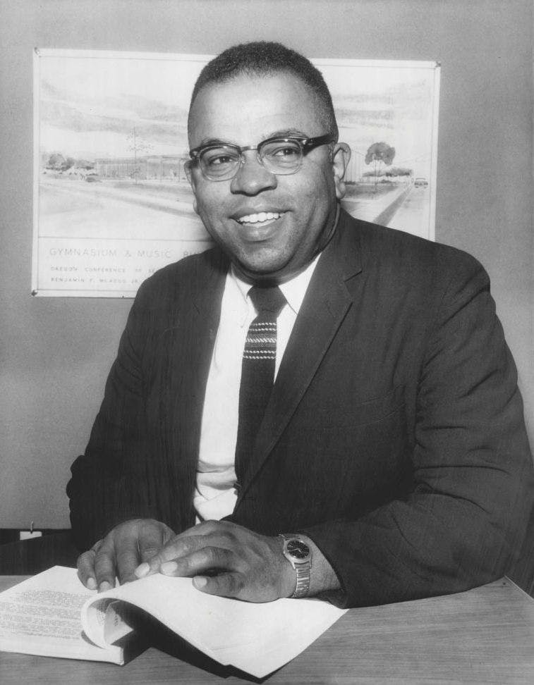 Person with glasses smiling, he's wearing a tie and jacket, his hands are on a book on table