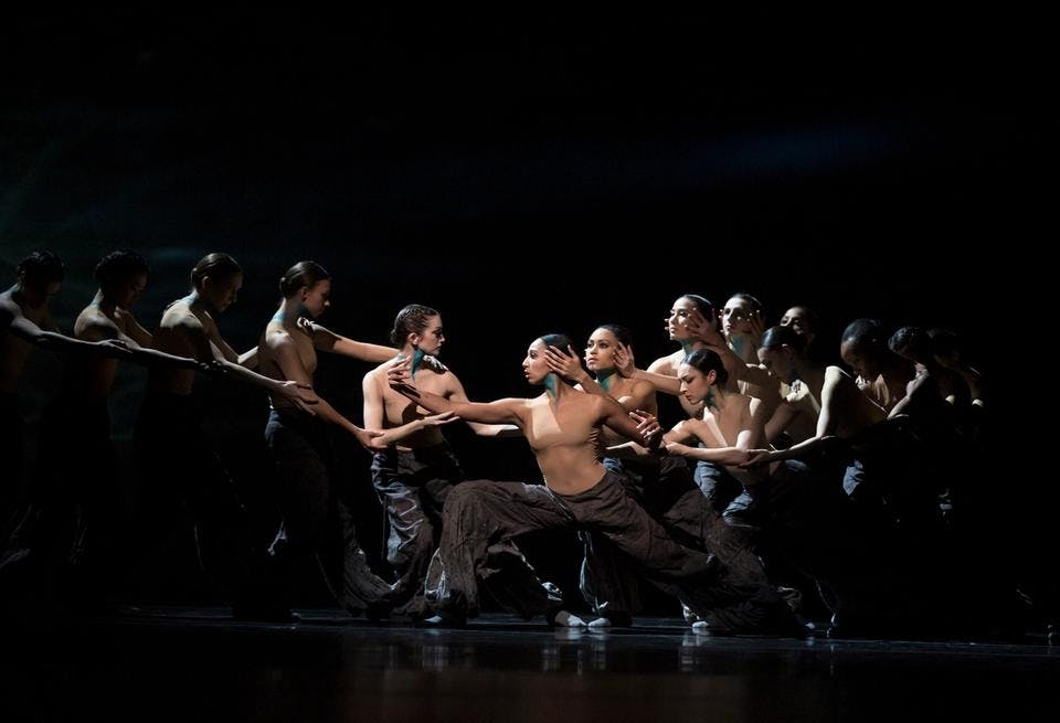 A large group of dancers in two rows lean toward each other on a dark stage