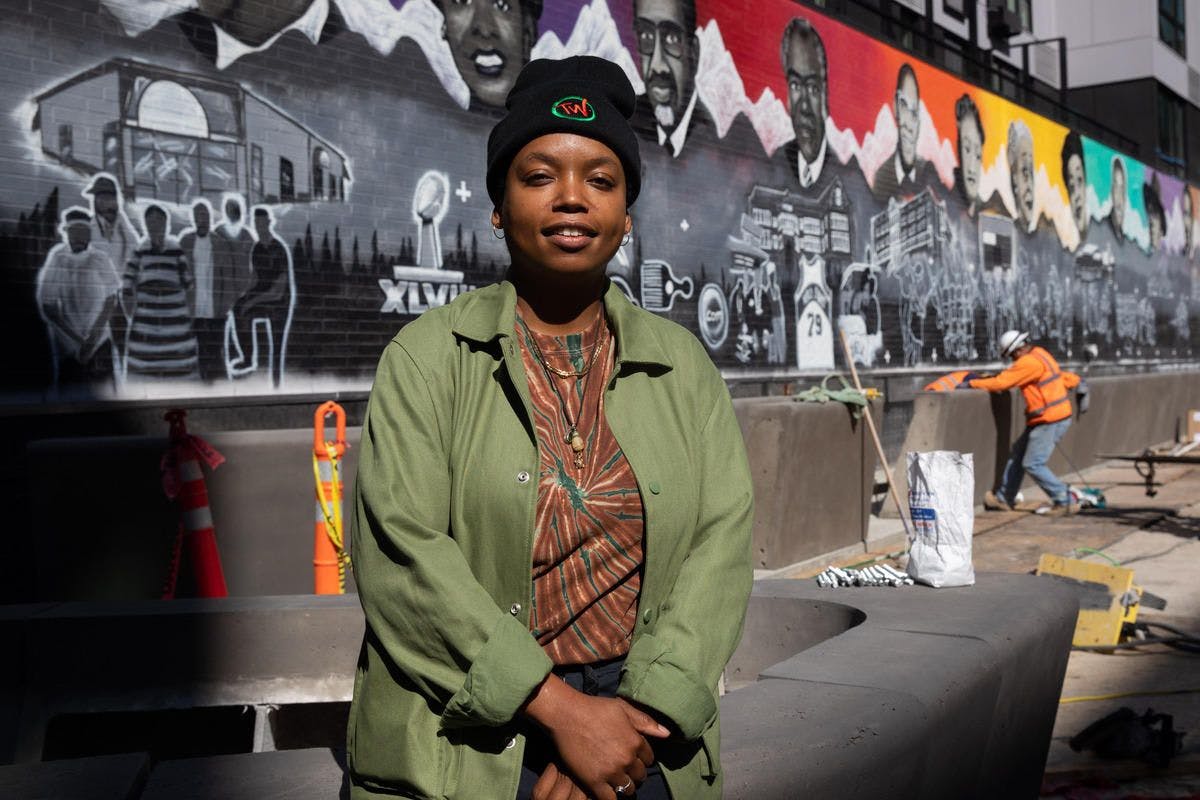 A woman in a green jacket and beanie stands in front of a mural