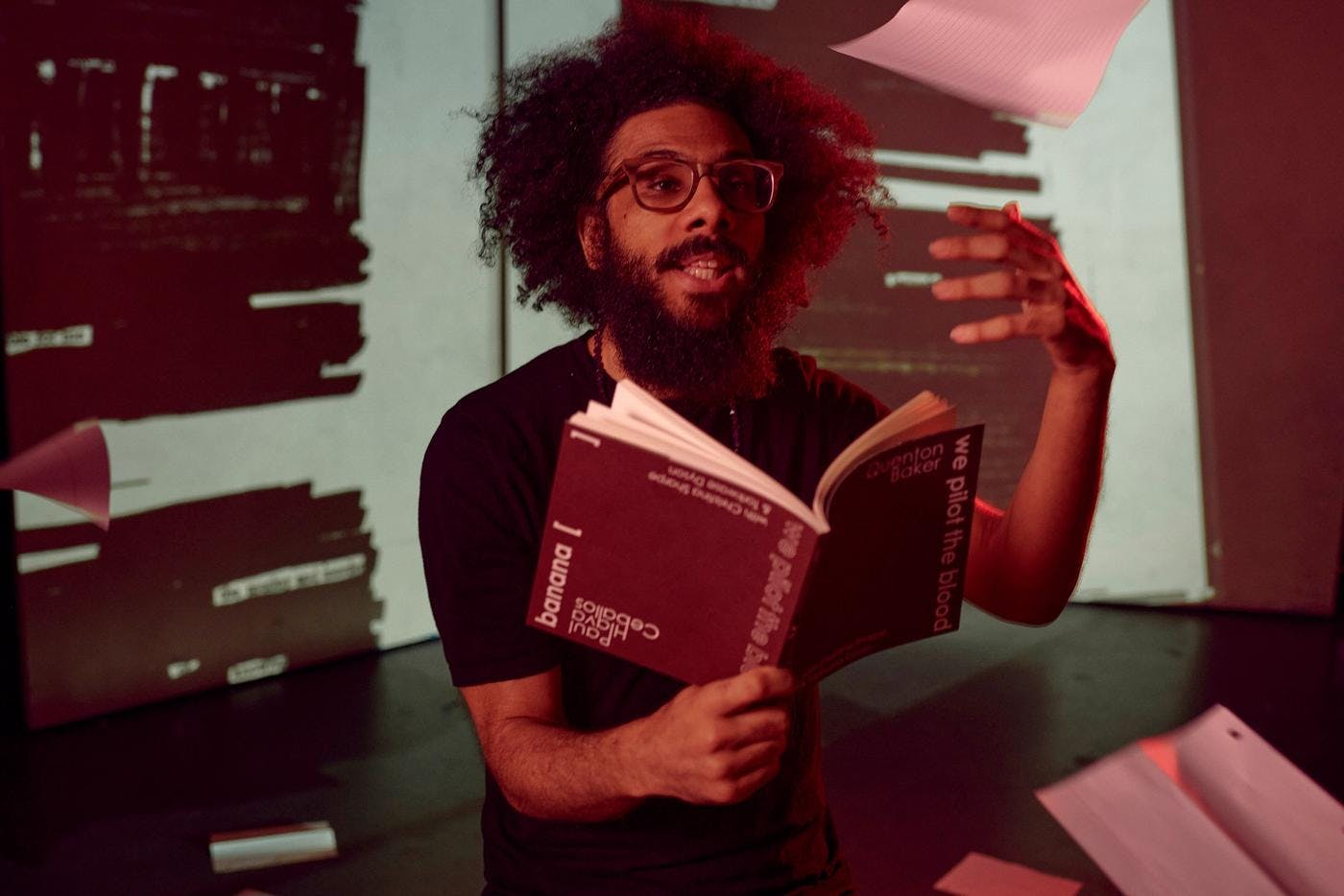 Quenton Baker reads from a book of poetry as sheets of notebook paper fall around him. A page from the book is projected on the a screen behind him.