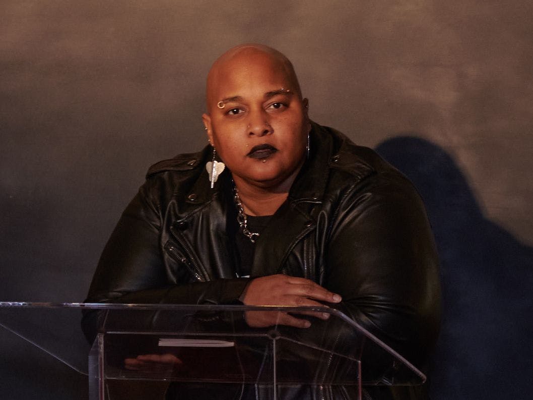 A bald woman wearing earrings and a leather jacket looks into the camera