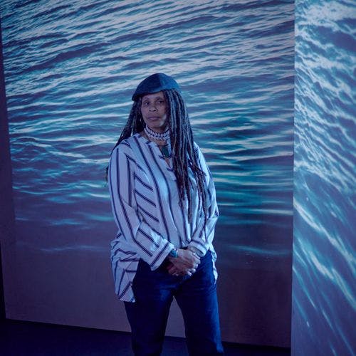 A woman stands in front of a projection of the ocean with her hands clasped