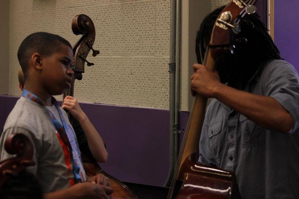 A boy tilts his head as he watches an instructor teach him how to play the cello