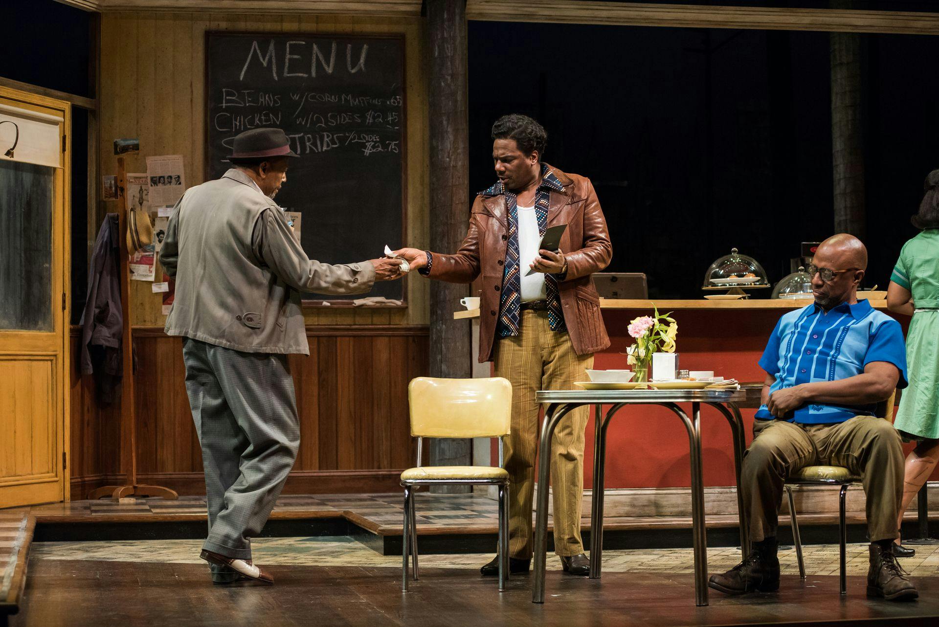In a play on stage, one man hands another cash, while a third man sits at a table