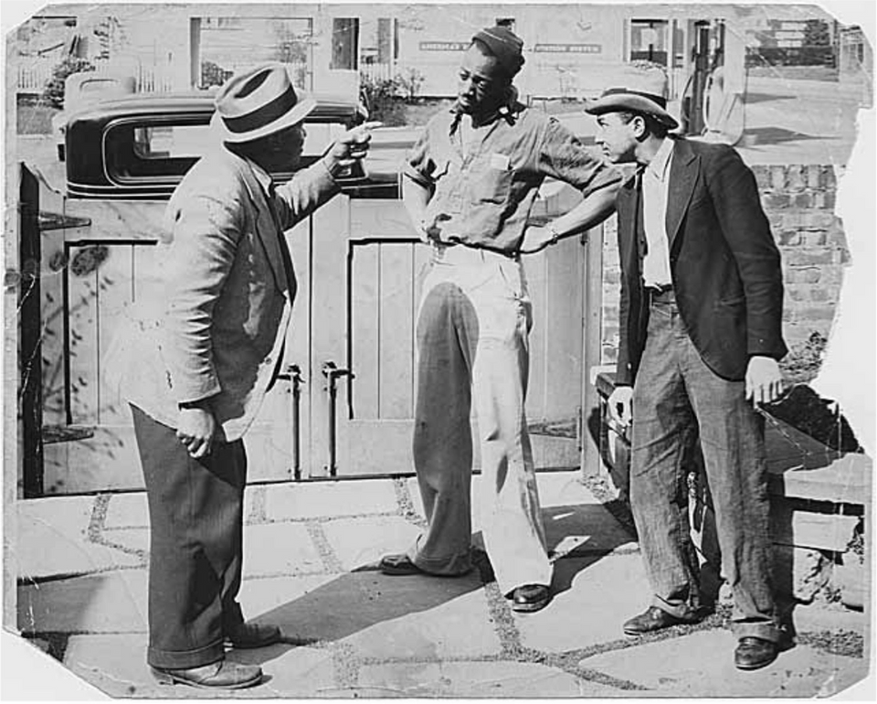 A black and white photo of a man yelling at another man, who has his hands on his hips, while another man looks on