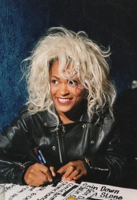 Person wearing black leather jacket smiles, their hair is platinum blonde. They are signing something on the table in front of them with a thick black marker.