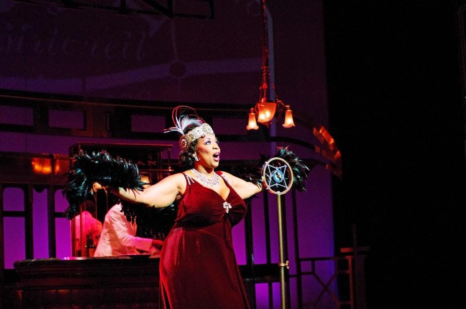 A stage scene with a woman in a red velvet dress holding feathers with arms outstretched